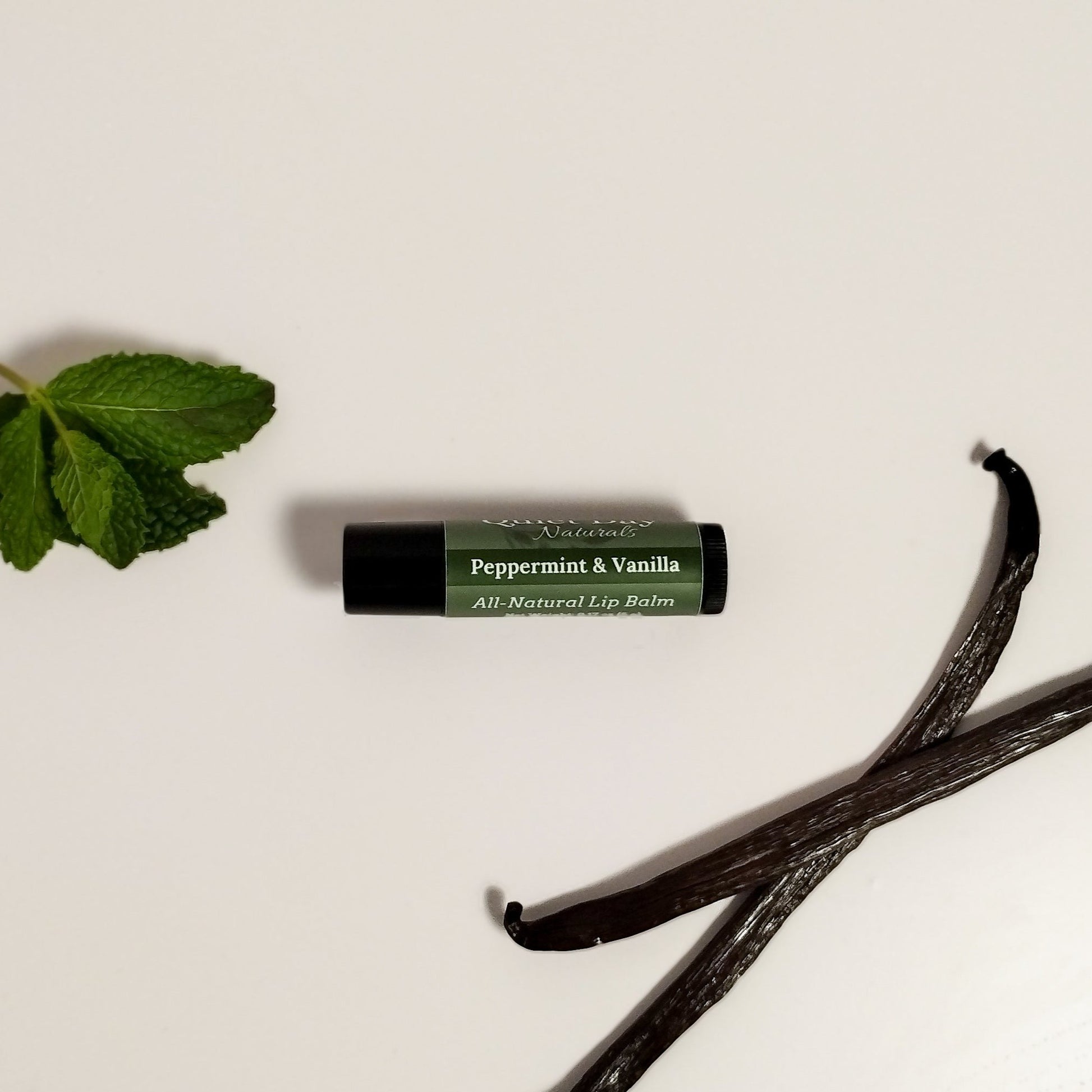 Handmade Natural Lip Balm laying on a white background with mint leaves and vanilla beans next to it.